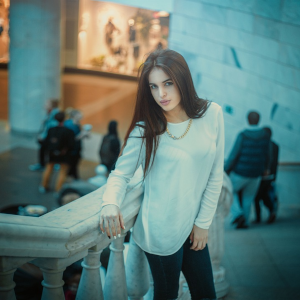 Молода красива дівчина з довгим темним волоссям у білій кофті, яка спирається на балюстраду в торговельному центрі.